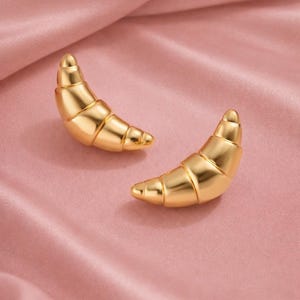 Puede incluir: Dos pendientes dorados con forma de croissant. Los pendientes están hechos de un material metálico brillante y se colocan sobre un fondo de satén rosa. Son una joya con temática de comida.