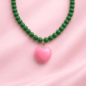 Puede incluir: Collar con cuentas verdes y un colgante de corazón rosa. El corazón cuelga de una argolla dorada. El collar se muestra sobre un fondo de satén rosa. Las cuentas tienen aproximadamente 0,5 cm de diámetro.