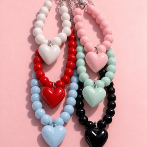 Puede incluir: Una colección de collares de cuentas con colgantes de corazón. Los collares presentan una variedad de colores, incluyendo blanco, rojo, azul, rosa, verde menta y negro. Cada collar tiene un colgante de corazón a juego.