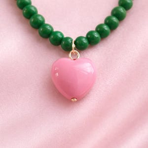 Puede incluir: Collar con un colgante de corazón rosa y cuentas verdes. El colgante de corazón es liso y brillante, suspendido de una fianza dorada. Las cuentas del collar son de un verde intenso, creando un contraste llamativo.
