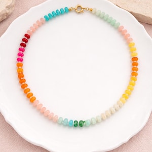 Puede incluir: Un collar de cuentas de colores con un degradado arcoíris de colores, incluyendo rojo, rosa, naranja, amarillo, verde y azul. El collar se muestra sobre un plato blanco sobre un fondo rosa. El cierre es dorado.