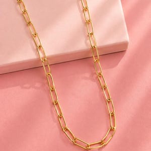 Puede incluir: Un collar de tono dorado que presenta una serie de eslabones alargados y rectangulares estilo clip. La cadena está construida con segmentos metálicos pulidos y uniformes conectados en un bucle continuo, creando un patrón geométrico repetitivo.