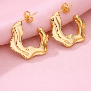 Puede incluir: Un par de pendientes de aro dorados de forma abstracta. Los pendientes tienen una apariencia texturizada de metal fundido y están sobre un fondo rosa suave. Los pendientes están diseñados con un cierre de poste.