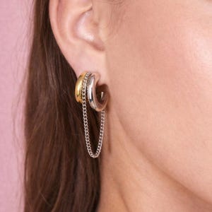 Puede incluir: Primer plano de un pendiente de aro plateado y dorado con una cadena. El pendiente tiene un aro plateado con un aro dorado y una cadena adjunta. El fondo es de color rosa suave.