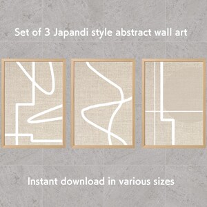 以下が含まれることがあります： 3枚セットの、Japandiスタイルの抽象的なウォールアートプリント。各プリントは、白い線形デザインが施されたベージュ色の背景です。フレームは明るい木の色です。上部のテキストは「Set of 3 Japandi style abstract wall art」と書かれています。