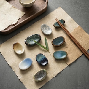 Può includere: Una collezione di piccoli poggiabacchette in ceramica di forma ovale in vari colori, tra cui blu, verde e beige. I poggiabacchette sono disposti su un pezzo di carta pergamena, con un paio di bacchette e una foglia di bambù. Un vassoio di legno e una piccola ciotola sono sullo sfondo.