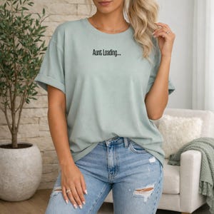 May include: A pale green t-shirt with the words "Aunt Loading..." printed in black. The shirt is worn with light blue jeans. The model is standing in front of a brick wall and a white chair. The sleeves are rolled up.
