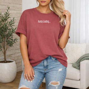 May include: A maroon t-shirt with the white text "Aunty Loading...". The top has a casual fit and short sleeves. The model is wearing blue jeans.