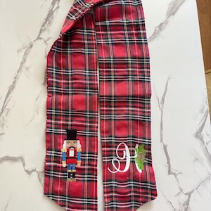 Puede incluir: Bufanda a cuadros rojos y negros con un cascanueces bordado en un extremo y una inicial blanca con acebo en el otro. Un accesorio festivo.