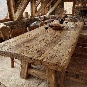 Op de afbeelding: Een grote, rustieke houten eettafel, ongeveer 300 cm lang, met een verweerde afwerking. Er staan wijnglazen en een broodmand op de tafel. De kamer heeft zichtbare houten balken en een stenen muur, wat een rustieke sfeer creëert.