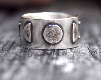 May include: A silver ring with a hammered texture and four moon phases. The ring is wide and has a textured surface.