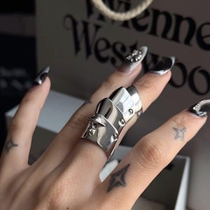 Peut inclure: Bague de doigt argentée avec un design en plusieurs parties, comprenant une série de plaques articulées qui recouvrent le doigt. La bague présente de petits détails de rivets et est portée sur une main avec du vernis à ongles noir et blanc et des tatouages.