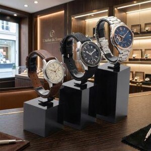 May include: Three wristwatches displayed on black pedestals. The watches feature various band styles, including brown leather, black rubber, and a silver metal bracelet. The watch faces showcase different designs and colors, including silver, black, and blue.