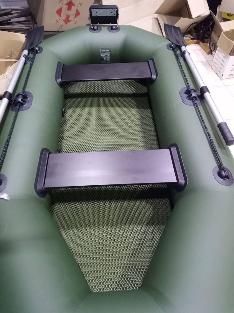 K&ouml;nnte beinhalten: Ein olivgr&uuml;nes Schlauchboot mit zwei schwarzen Sitzb&auml;nken, einem strukturierten olivgr&uuml;nen Boden und zwei silbernen Rudern mit schwarzen Bl&auml;ttern an den Seiten. Eine schwarze Motorhalterung befindet sich am Heck des Bootes.
