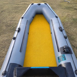 K&ouml;nnte beinhalten: Ein Schlauchboot mit hellgrauer Au&szlig;enh&uuml;lle, schwarzen Verzierungen und einem leuchtend gelben, strukturierten Boden. Das Boot verf&uuml;gt &uuml;ber schwarze Griffe, Seilf&uuml;hrungen und ein kleines rechteckiges Etikett mit blau-wei&szlig;em Text auf der rechten Seite.