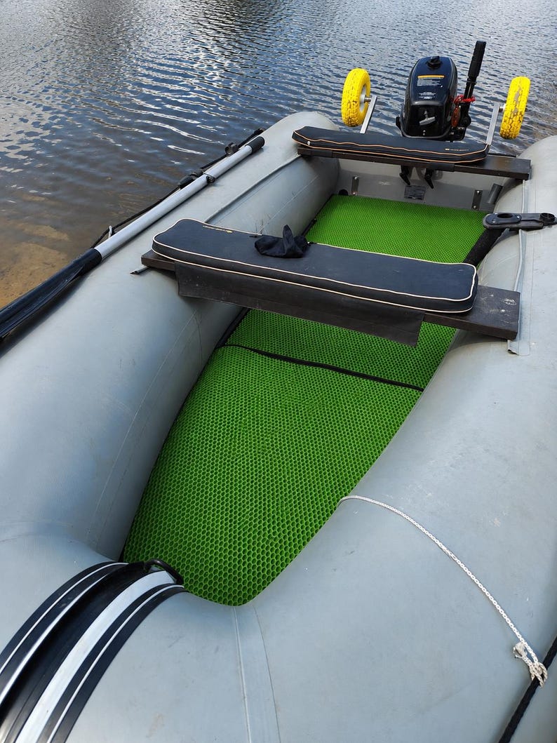 K&ouml;nnte beinhalten: Ein aufblasbares graues Gummiboot mit einer leuchtend gr&uuml;nen Bodenmatte im Wabenmuster, zwei schwarzen Sitzb&auml;nken mit wei&szlig;er Kante und einem kleinen Au&szlig;enbordmotor am Heck, der mit zwei gelben R&auml;dern f&uuml;r den Transport ausgestattet ist.