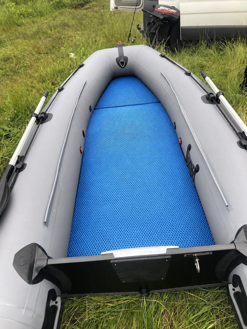 K&ouml;nnte beinhalten: Ein Schlauchboot mit hellgrauen Schl&auml;uchen und einer leuchtend blauen, strukturierten Bodenmatte. Das Boot verf&uuml;gt &uuml;ber schwarze Ruderdollen, seitliche Halteleinen und einen schwarzen Heckspiegel. Der Boden ist mit einer blauen Anti-Rutsch-Matte ausgelegt.