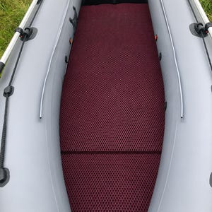 K&ouml;nnte beinhalten: Ein Schlauchboot mit hellgrauen Seitenschl&auml;uchen und einer dunkelroten Bodenmatte mit Rautenmuster. Der Innenraum verf&uuml;gt &uuml;ber schwarze Seitenw&auml;nde, schwarze Ruderdollen und eine schwarze Heckplatte mit kleinen wei&szlig;en Warnaufklebern.