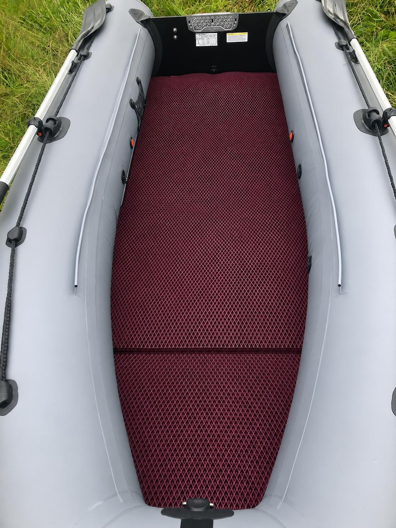K&ouml;nnte beinhalten: Ein Schlauchboot mit hellgrauen Seitenschl&auml;uchen und einer dunkelroten Bodenmatte mit Rautenmuster. Der Innenraum verf&uuml;gt &uuml;ber schwarze Seitenw&auml;nde, schwarze Ruderdollen und eine schwarze Heckplatte mit kleinen wei&szlig;en Warnaufklebern.