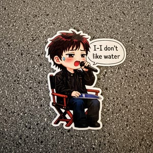Puede incluir: Una pegatina con un personaje de estilo anime sentado en una silla de director, sosteniendo un micrófono. El personaje tiene el pelo rojo, una chaqueta negra y vaqueros azules. Un bocadillo dice: "I-I don't like water."