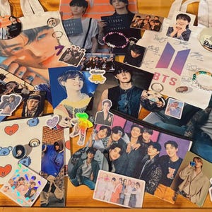 May include: A display of BTS merchandise, such as tote bags, stickers, bracelets, and photo cards, on a wooden surface. The items showcase images of the band members and the BTS logo. The colour scheme includes white, purple, and various skin tones.
