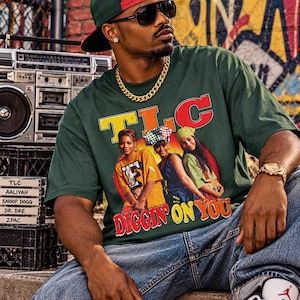 May include: A dark green short-sleeve t-shirt featuring a graphic print of the music group TLC with the text TLC and DIGGIN ON YOU. The shirt is worn with blue denim jeans, a gold chain necklace, a green baseball cap, and dark sunglasses.
