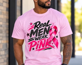 Puede incluir: Una camiseta de manga corta de color rosa claro con un diseño gráfico que incluye el texto Real MEN WEAR PINK en letras negras y rosas estilo pincelada, acompañado de un lazo rosa de concienciación y pequeñas estrellas negras al frente.