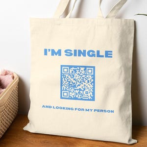 Może przedstawiać: Kremowa torba typu tote z niebieskim napisem "I'M SINGLE" nad niebieskim kodem QR i napisem "AND LOOKING FOR MY PERSON". Torba ma długie uchwyty i jest wykonana z materiału przypominającego płótno.