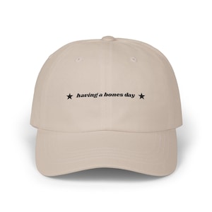 Könnte beinhalten: Beige Baseballkappe mit gebogenem Schirm. Der Aufdruck "having a bones day" in schwarzer Schrift, mit einem schwarzen Stern auf jeder Seite. Die Kappe ist aus einem weichen Stoff gefertigt.