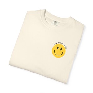 以下が含まれることがあります： クリーム色のTシャツ。黄色いスマイリーフェイスのグラフィックと「new mom who dis」の文字がプリントされています。シャツは折りたたまれており、襟にはタグが付いています。スマイリーフェイスは黒い目と曲がった口をしています。