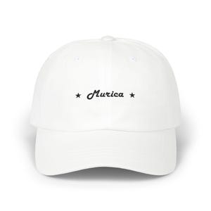 Puede incluir: Gorra de béisbol blanca con visera curva. La gorra presenta la palabra "Murica" en una fuente cursiva, flanqueada por estrellas negras. El sombrero está hecho de un material suave y tiene una correa ajustable.