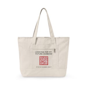 May include: Cream-colored tote bag with a front pocket and long handles. The bag features the text "LOOKING FOR MY FUTURE HUSBAND" above a red QR code. Below the code, the text reads "IF YOU FELT THE ENERGY, SCAN IT."