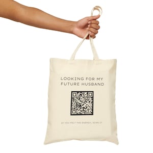 Op de afbeelding: Crèmekleurige draagtas met de tekst "LOOKING FOR MY FUTURE HUSBAND" boven een QR-code. Daaronder staat "IF YOU FELT THE ENERGY, SCAN IT". De tas wordt door een persoon vastgehouden.