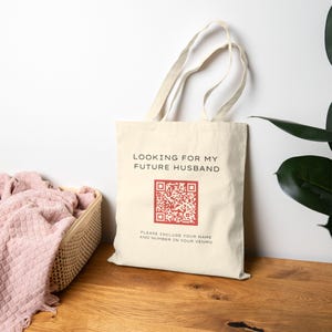 May include: Cream-colored tote bag with the text "LOOKING FOR MY FUTURE HUSBAND" above a red QR code. Below the QR code is the text "PLEASE INCLUDE YOUR NAME AND NUMBER IN YOUR VENMO". The bag has a simple design and long handles.