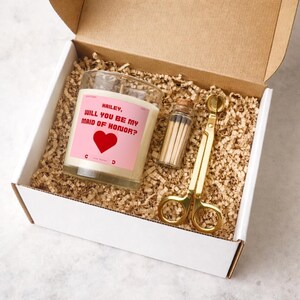 Könnte beinhalten: Geschenkbox mit Kerze, Streichhölzern und Dochtschere. Die Kerze hat ein rosa Etikett mit dem Text "Hailey, Will You Be My Maid of Honor?" und einem roten Herz. Die Box ist mit Papier gefüllt.