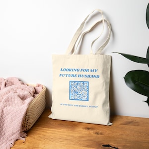 Puede incluir: Un bolso de tela color crema con dos asas para el hombro. La parte frontal presenta texto en azul claro que dice "LOOKING FOR MY FUTURE HUSBAND" sobre un código QR azul claro, seguido del texto "IF YOU FELT THE ENERGY, SCAN IT" debajo.