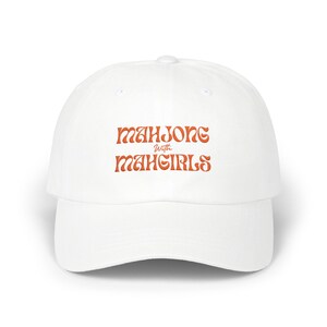 May include: White baseball cap with the text "Mahjong with Mahgirls" in orange lettering. The cap has a curved brim and a rounded crown. A casual accessory for mahjong enthusiasts.