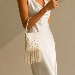 May include: A white satin slip dress with spaghetti straps. A small, rectangular, off-white handbag with pearl embellishments and a fringe of hanging pearls is held by the dress's wearer. The dress and bag suggest a formal or bridal theme.