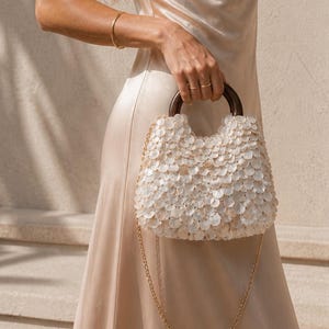 Könnte beinhalten: Cremefarbenes Satin-Slip-Kleid mit Wasserfallausschnitt. Eine kleine, weiße, paillettenbesetzte Handtasche mit Holzgriff und goldener Kette wird gehalten. Der Hintergrund ist eine neutrale Wand mit Schatten.