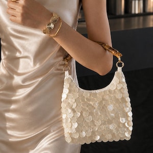 May include: A cream-coloured handbag with a scalloped edge, covered in round, iridescent sequins. The bag has a bamboo handle and is held by a person wearing gold jewellery, including a ring and bracelet. The person is wearing a satin dress.