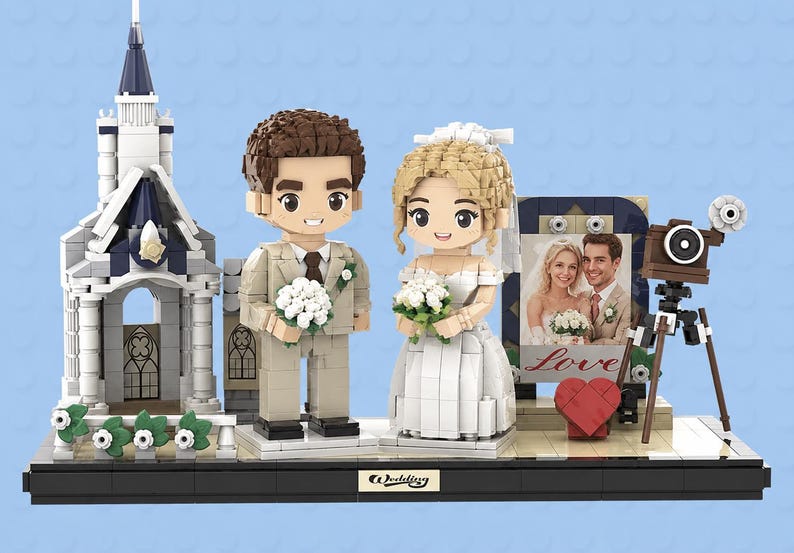 May include: A building block set featuring a bride and groom figurine, a white church structure, a vintage-style camera on a tripod, and a framed photograph of a couple with the word Love printed below. The base has a plaque labeled Wedding.