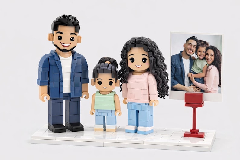 May include: A custom brick-built family figurine set featuring three figures standing on a white base next to a small red stand displaying a square photograph of the same family. The figures have detailed hair, facial features, and colorful clothing.
