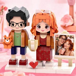 May include: A building block set featuring two stylized figures standing on a white base. The left figure has black hair, glasses, a green shirt, and a brown jacket, holding a small orange bear. The right figure has orange hair, a red dress, and a brown jacket, holding a drink. A photo frame displays a picture of a couple.