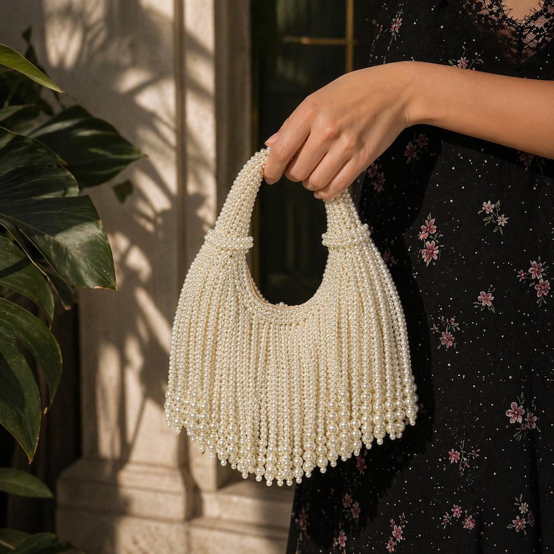 May include: A cream-colored handbag made of numerous strands of faux pearls. The bag has a rounded shape with a handle and a fringe-like bottom. The bag is held by a person.