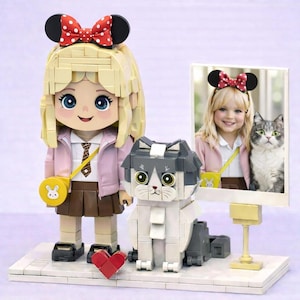 May include: A building block figure of a blonde character with a red polka-dot bow, a pink jacket, and a brown skirt, standing next to a block-built gray and white cat and a framed photograph of a child with a cat on a white base.