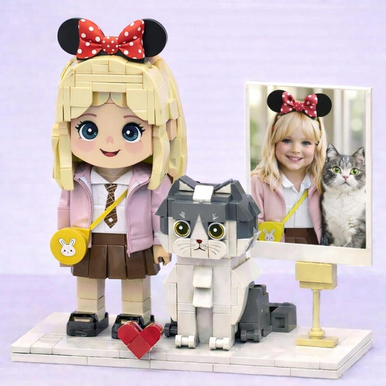 May include: A building block figure of a blonde character with a red polka-dot bow, a pink jacket, and a brown skirt, standing next to a block-built gray and white cat and a framed photograph of a child with a cat on a white base.