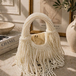 May include: A cream-colored handbag with a rounded handle and a fringe of faux pearls. The bag's body is also covered in pearls, creating a luxurious texture. The interior is a light beige color. A book titled "CHANEL THE VOCABULARY OF STYLE" is in the background.