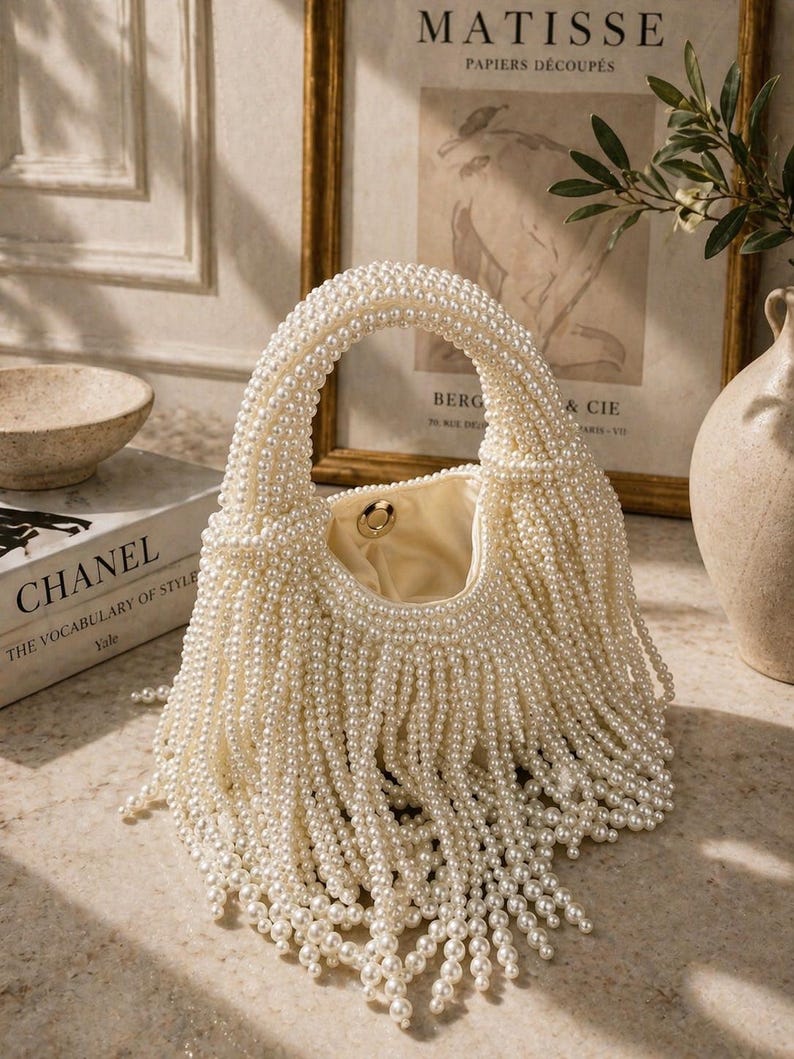 May include: A cream-colored handbag with a rounded handle and a fringe of faux pearls. The bag's body is also covered in pearls, creating a luxurious texture. The interior is a light beige color. A book titled "CHANEL THE VOCABULARY OF STYLE" is in the background.