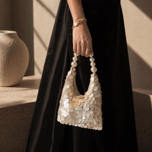 May include: A cream-colored sequined handbag with a pearl handle is held by a person wearing a gold bracelet and ring. The bag is made of small, round, iridescent sequins. The background features a black dress and a neutral-toned wall.