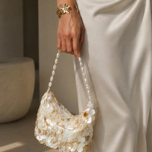 May include: A small handbag covered in overlapping white and iridescent shell-like discs, featuring a handle made of white oval beads. The bag is paired with a gold-toned metal flower bracelet and a smooth, light-colored satin fabric garment.
